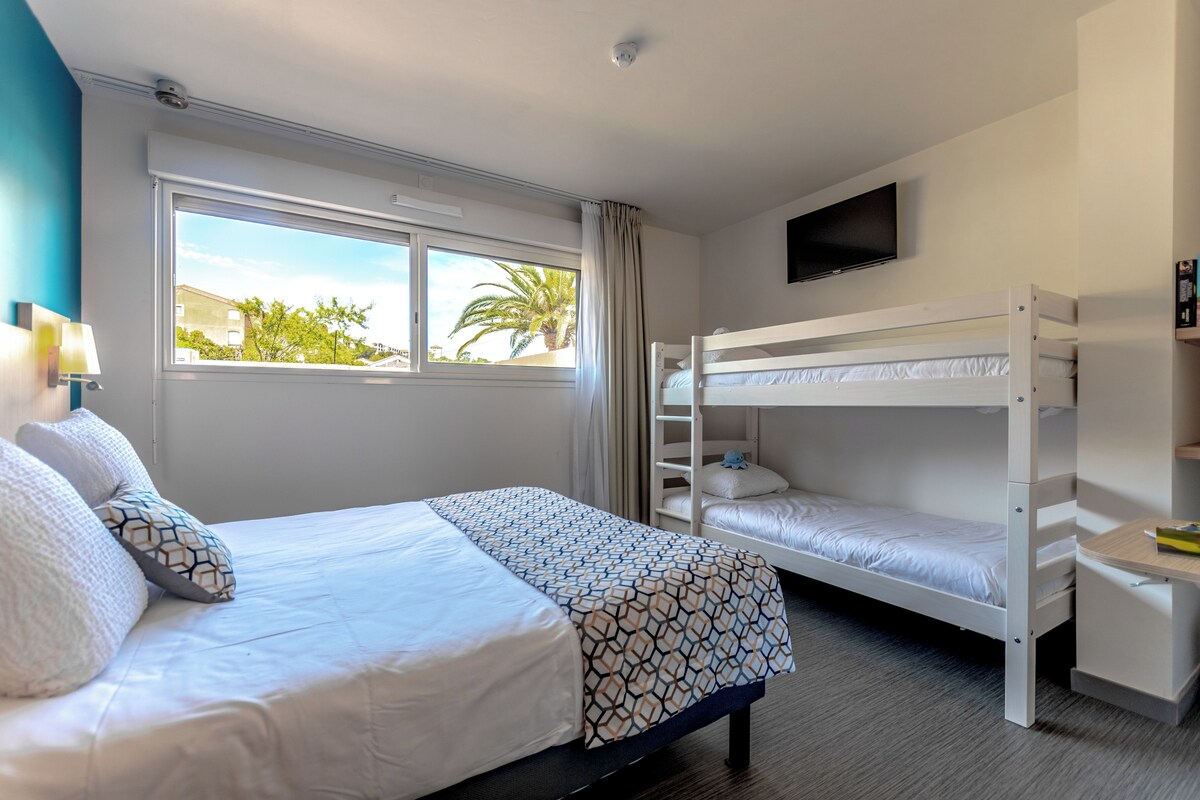 A hotel room with a bed, a bunk bed, a desk, a TV, and a window with a view of palm trees.