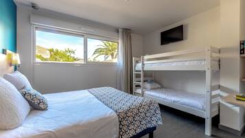 Habitación de hotel con cama, literas, escritorio, televisor y ventana con vista a palmeras.