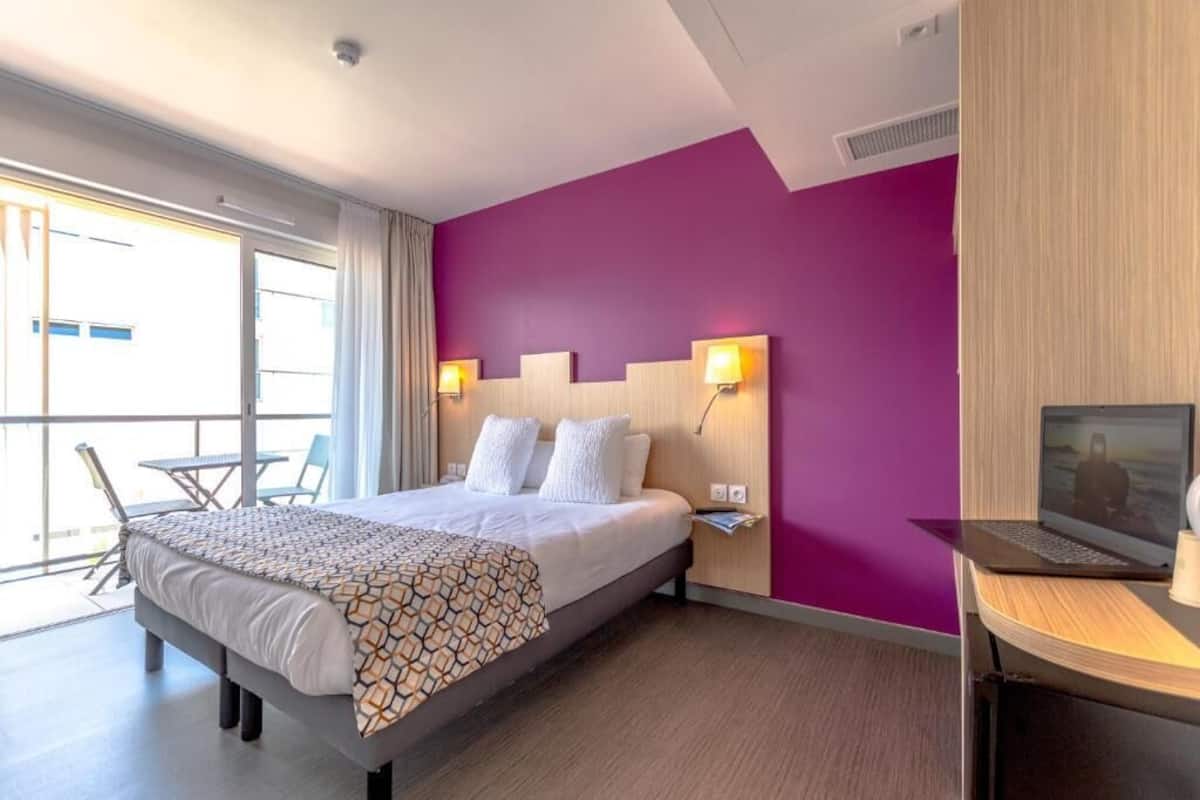A hotel room with a large bed, a desk with a laptop, and a view of the outdoors.