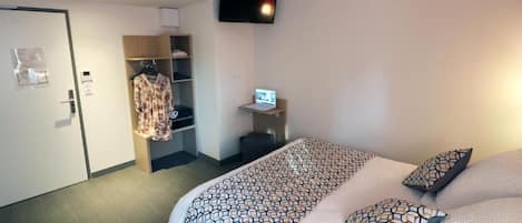 A hotel room with a bed, a TV mounted on the wall, a wardrobe, and a desk with a computer.