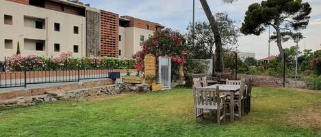 Zona residencial con césped, una mesa y sillas de madera, y un edificio con balcones y una pared de piedra.