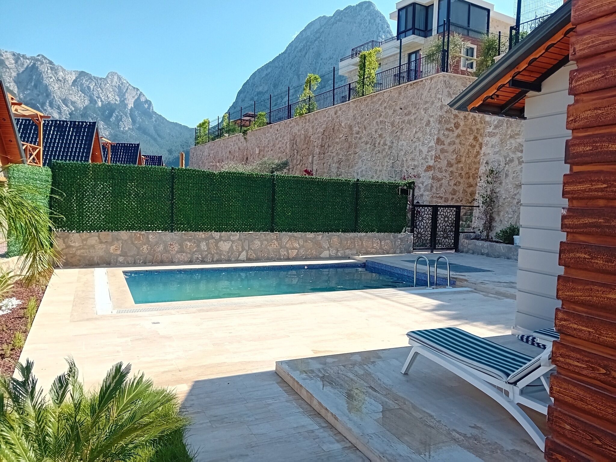 A swimming pool with a stone wall and a wooden deck, surrounded by greenery and a mountain backdrop.