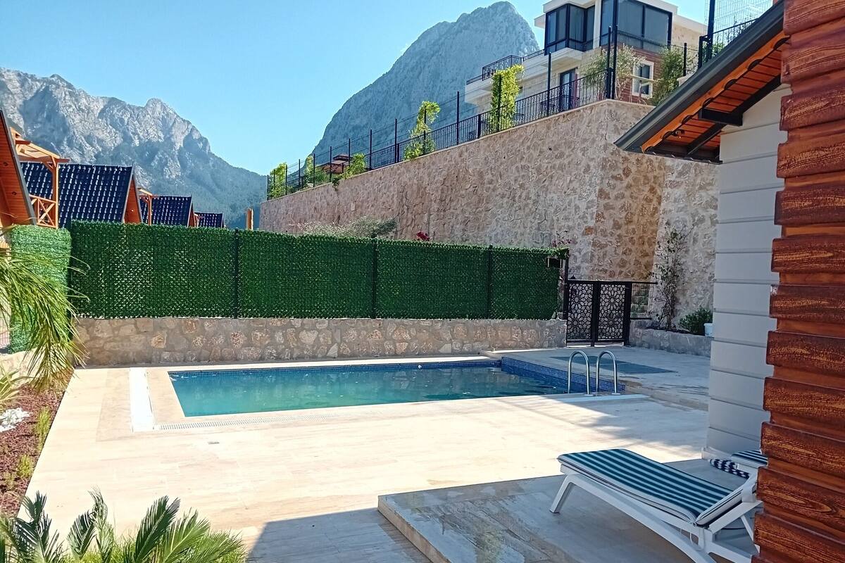 A swimming pool with a stone wall and a wooden deck, surrounded by greenery and a mountain backdrop.