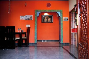 Een kamer met een oranje muur, een lijst met afbeeldingen van godheden, een klok en een deur met een decoratief patroon.