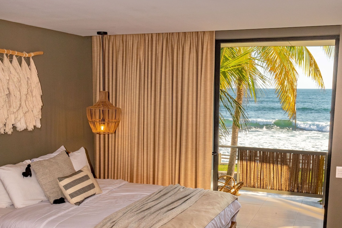 A bedroom with a bed, a potted plant, a hanging light fixture, and a view of the ocean.