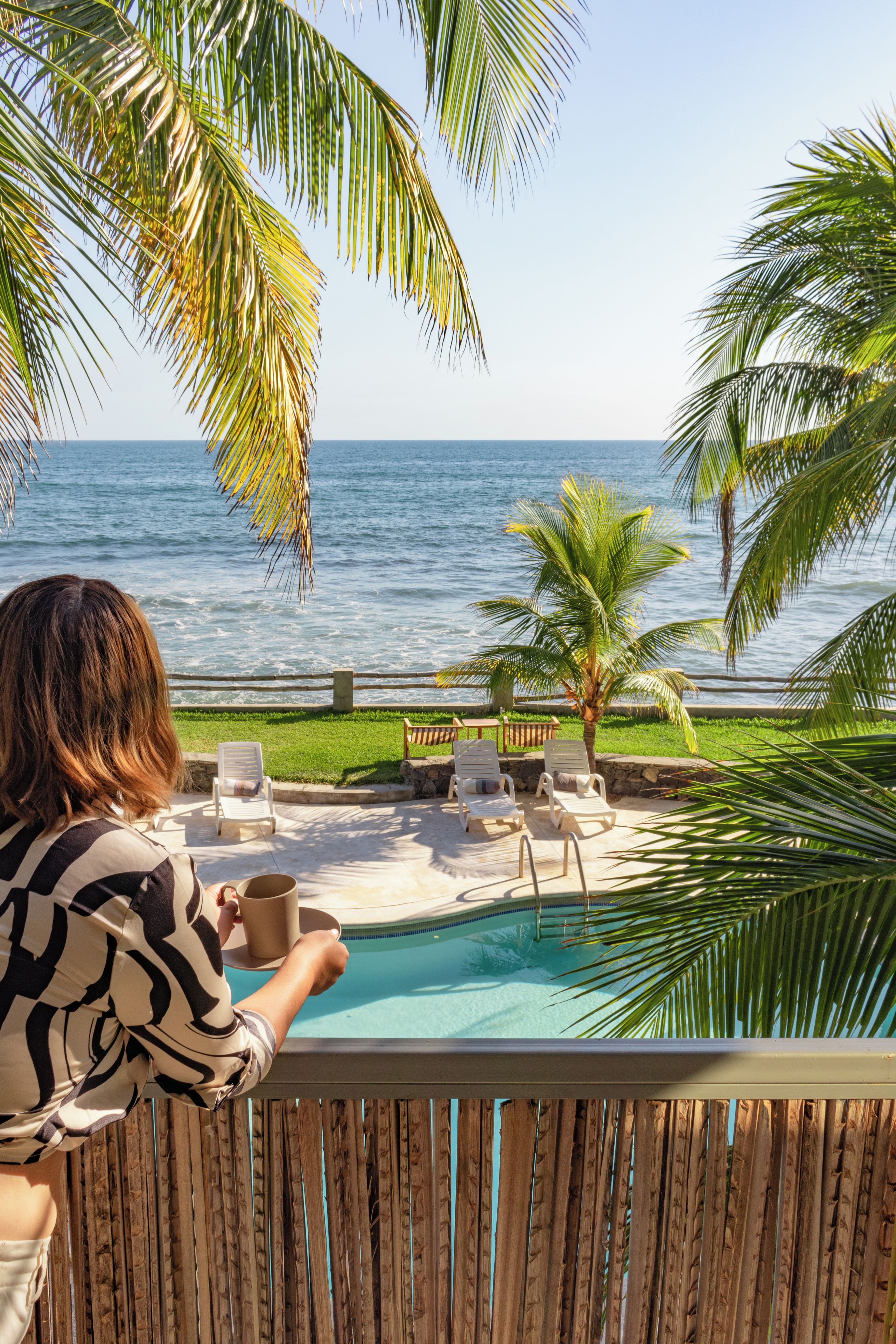 A poolside area with lounge chairs, a palm tree, and a view of the ocean.