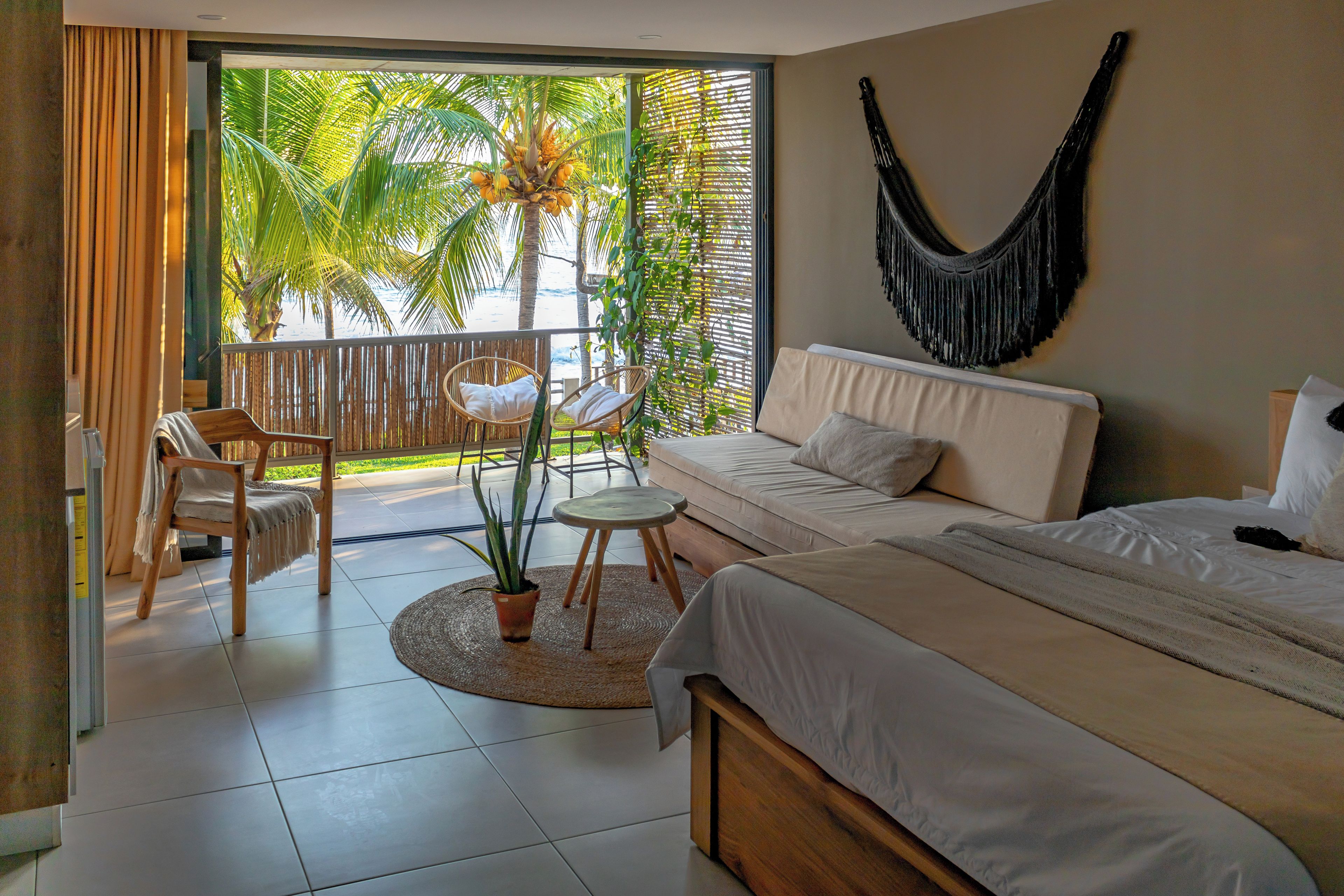 Ein Schlafzimmer mit Bett, Sofa, Sessel, einem kleinen Tisch, einer Topfpflanze und Blick auf Palmen vom Balkon aus.