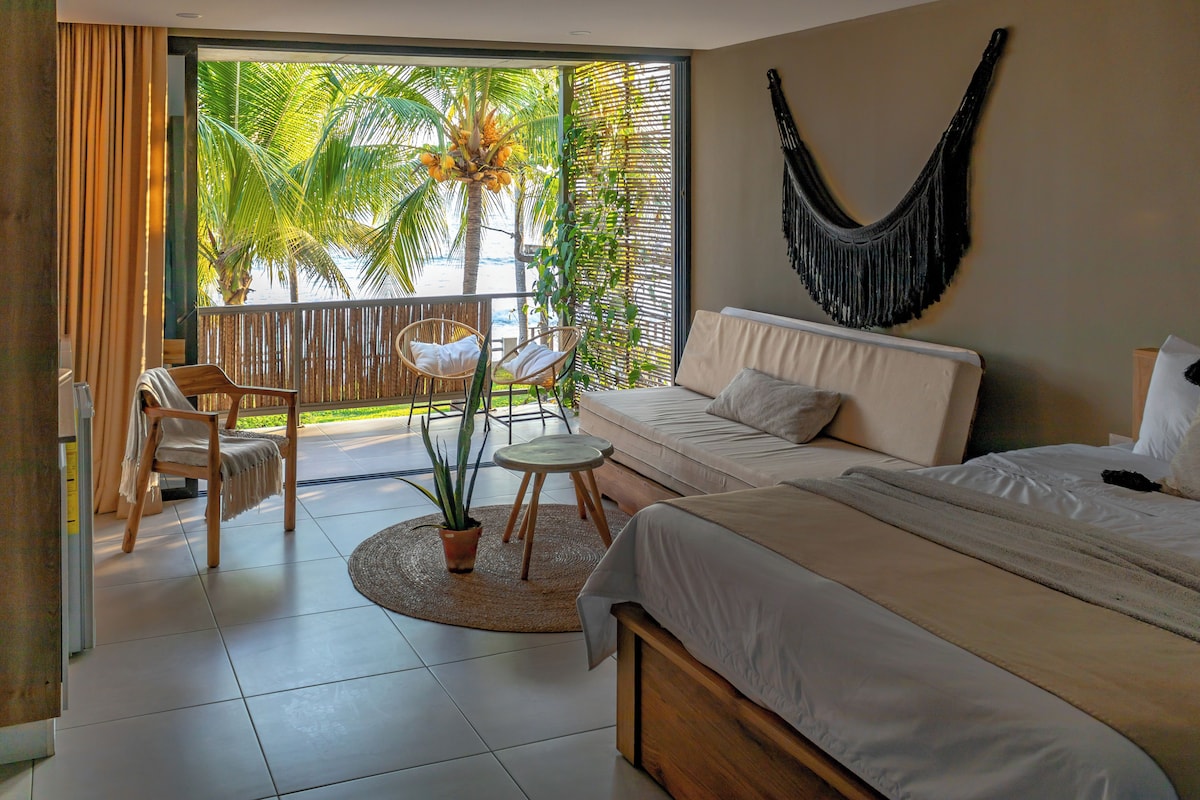 Un dormitorio con cama, sofá, sillón, una mesita, planta en maceta y vistas a palmeras desde un balcón.