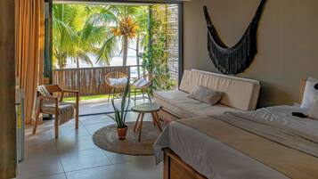 Ein Schlafzimmer mit Bett, Sofa, Sessel, einem kleinen Tisch, einer Topfpflanze und Blick auf Palmen vom Balkon aus.