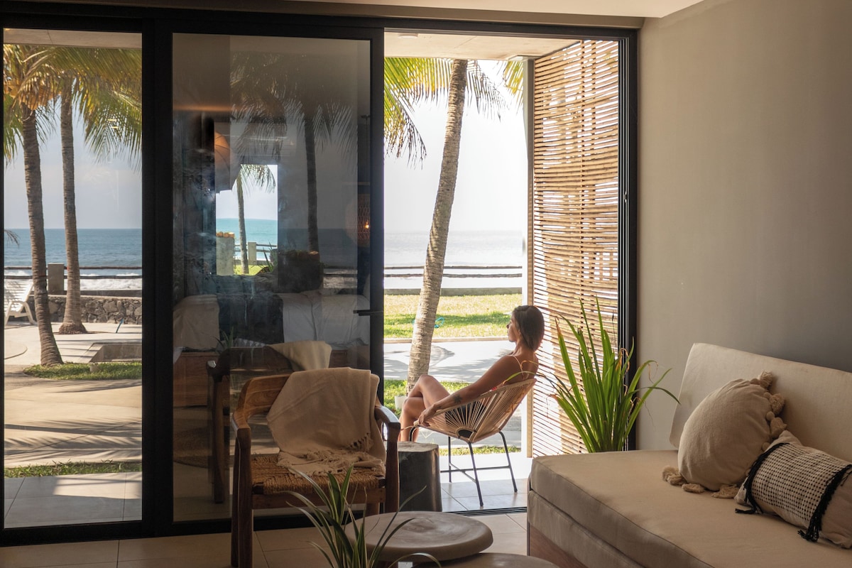 Una habitación con un amplio ventanal corredera que da a vistas de la playa, un sofá, una silla y una mesita.