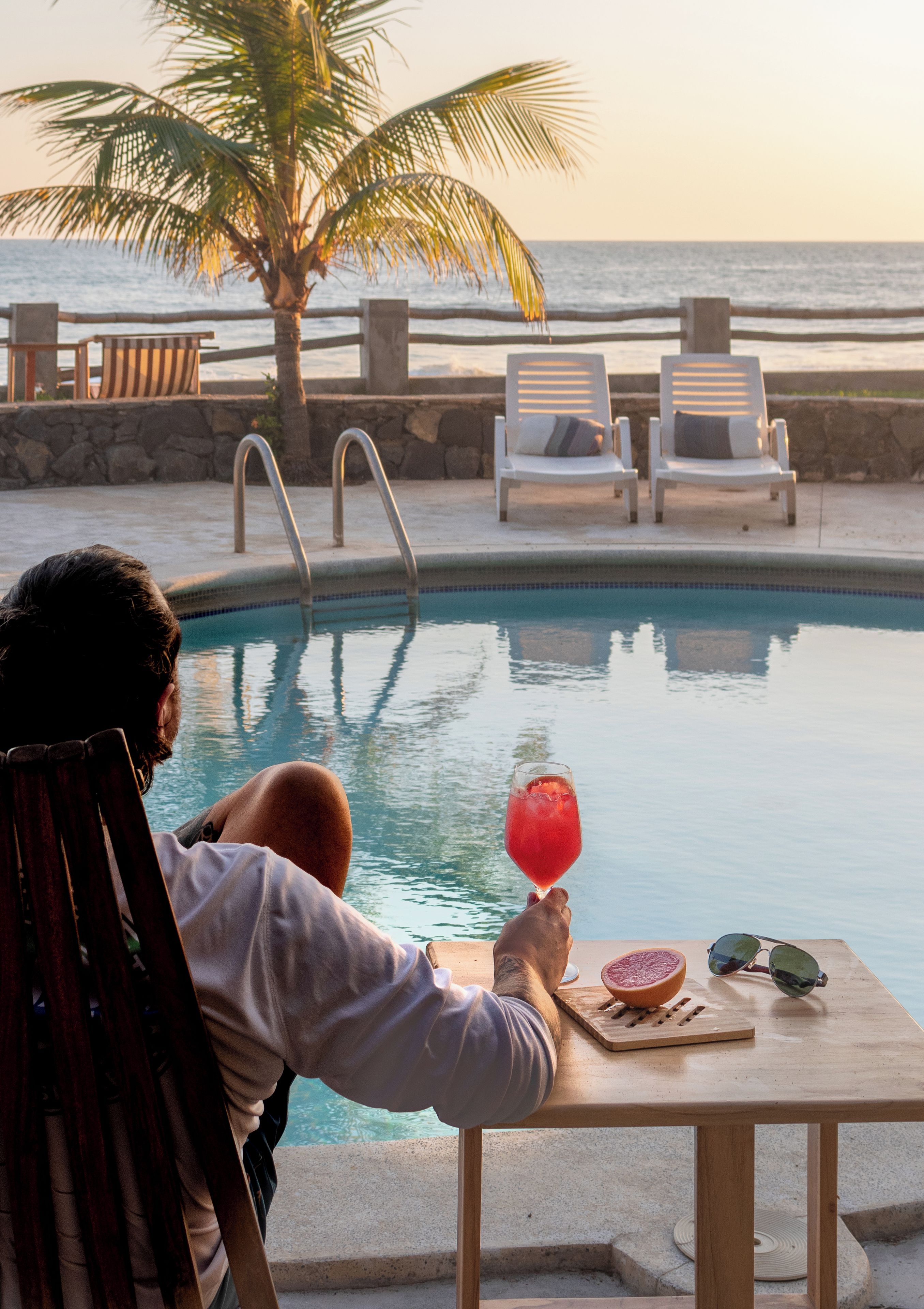A poolside setting with a person holding a drink, a table with a fruit, and lounge chairs.
