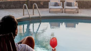 A poolside setting with a person holding a drink, a table with a fruit, and lounge chairs.