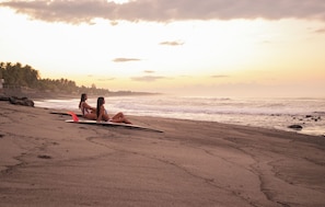 Una playa con dos personas sentadas en la arena, una tabla de surf y un mar tranquilo.