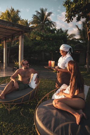 Uma pessoa segurando uma bandeja com bebidas, sentada em uma cadeira estofada ao ar livre, com palmeiras ao fundo.
