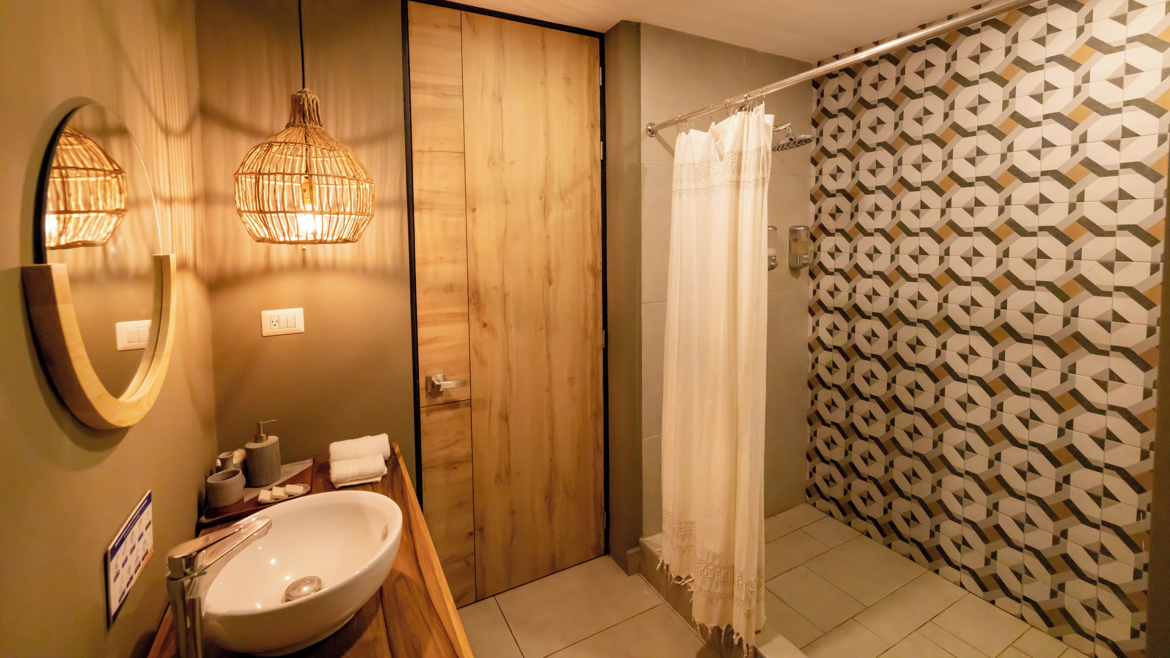 Ein Badezimmer mit Holztür, Waschbecken, Spiegel, einer Pendelleuchte und einer Dusche mit gemusterten Fliesen.
