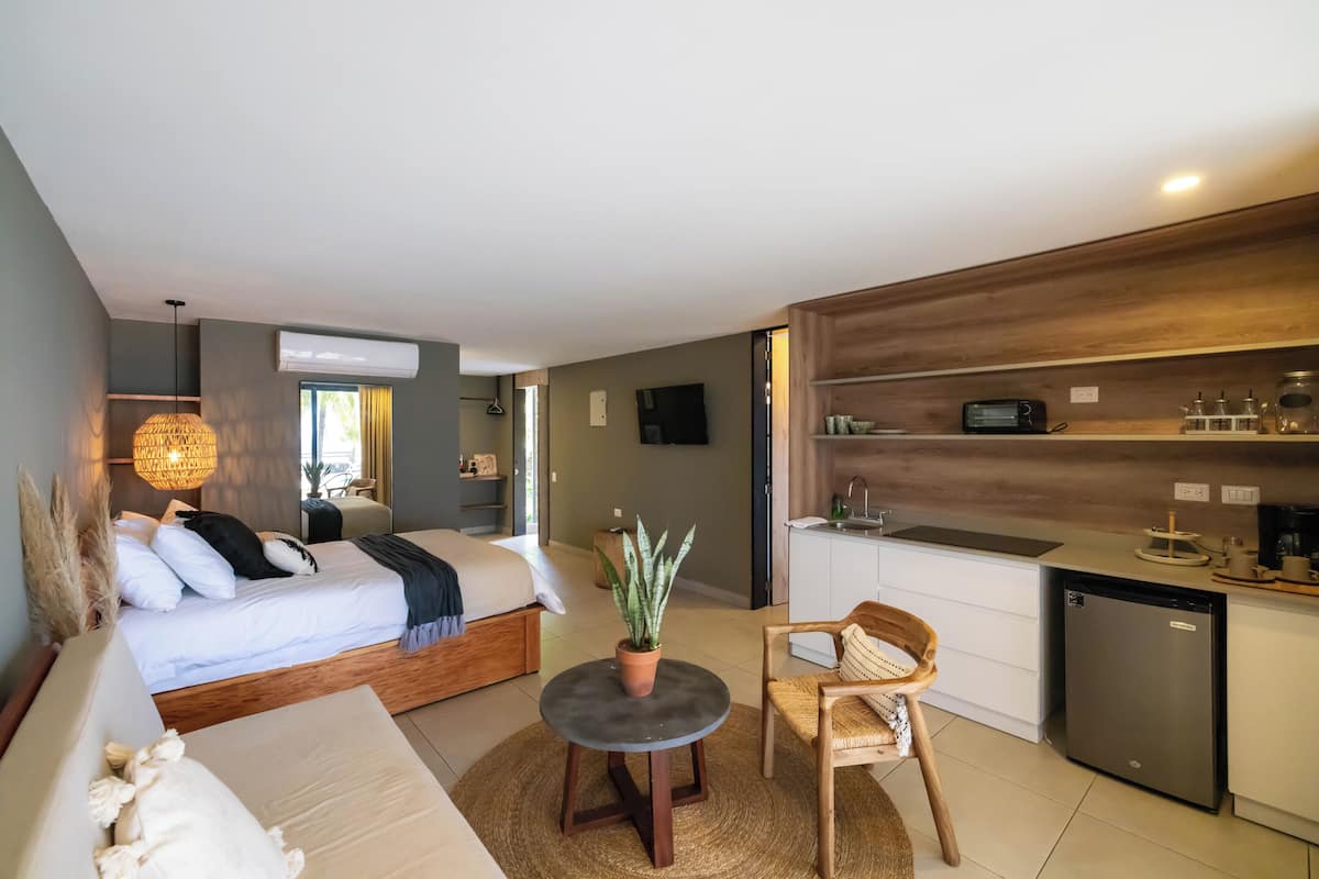 Ein modernes Hotelzimmer mit einem Bett, einem kleinen Esstisch mit Stuhl, einer Küchenzeile und einer Topfpflanze.
