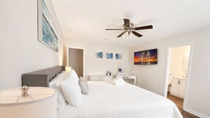 Ein Schlafzimmer mit Bett, Nachttischen, Deckenventilator, Kunstwerken und einem durchgegangenen Tür sichtbar liegenden Badezimmer.
