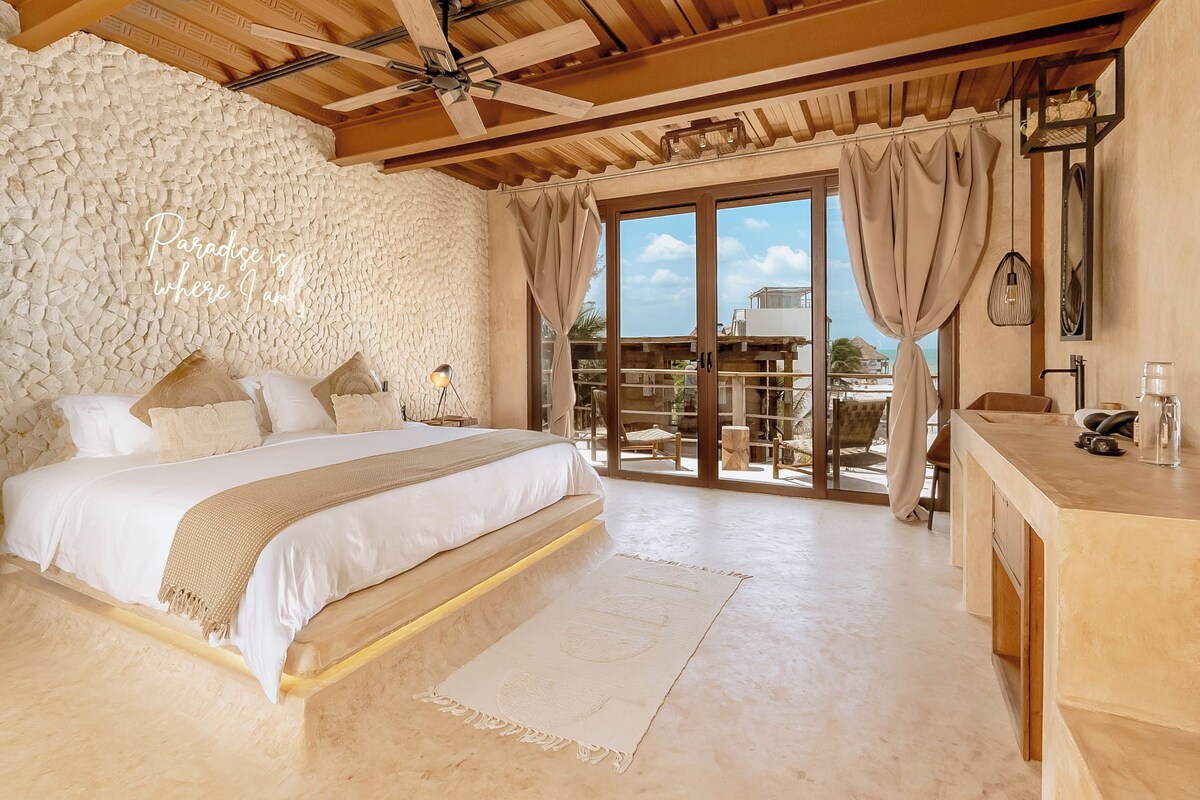 Een slaapkamer met een stenen muur, een bed met wit beddengoed, een balkon met gordijnen en uitzicht op zee.