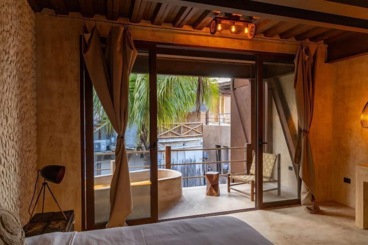A modern room with a bed, a small table, a chair, and a view of a pool and palm trees.