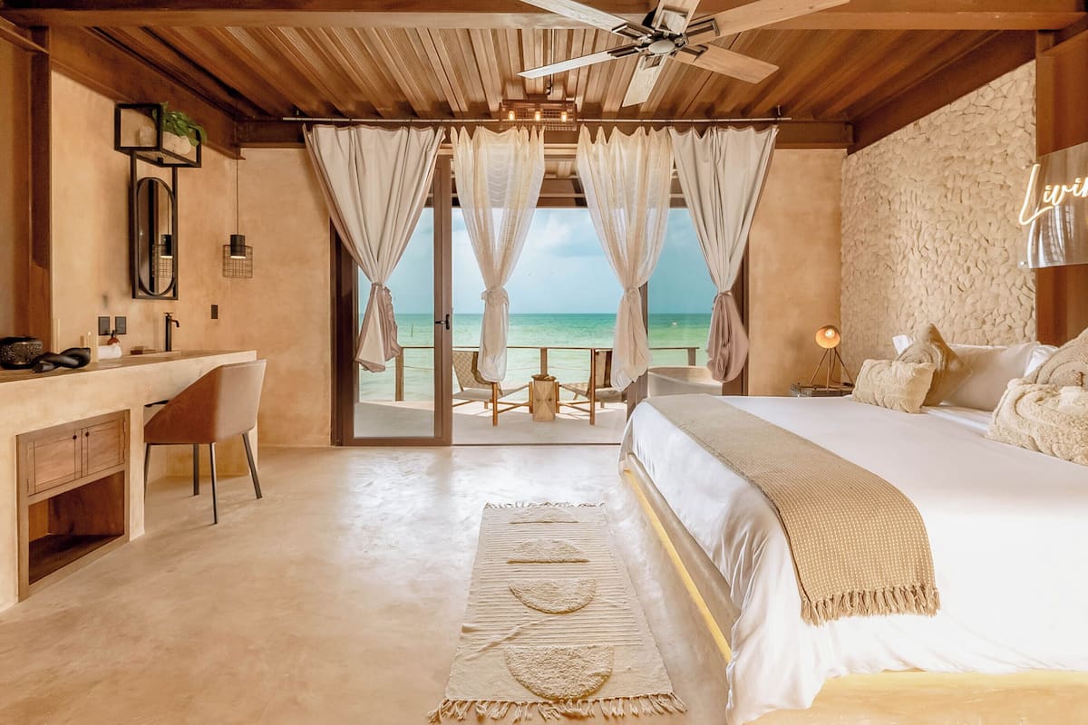A bedroom with a large bed, a desk, and a chair, with a view of the ocean through sheer curtains.