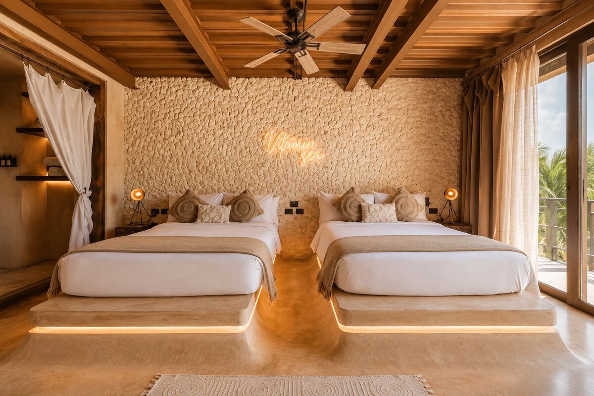 A double room with two beds, a stone wall, a ceiling fan, and a view of palm trees.