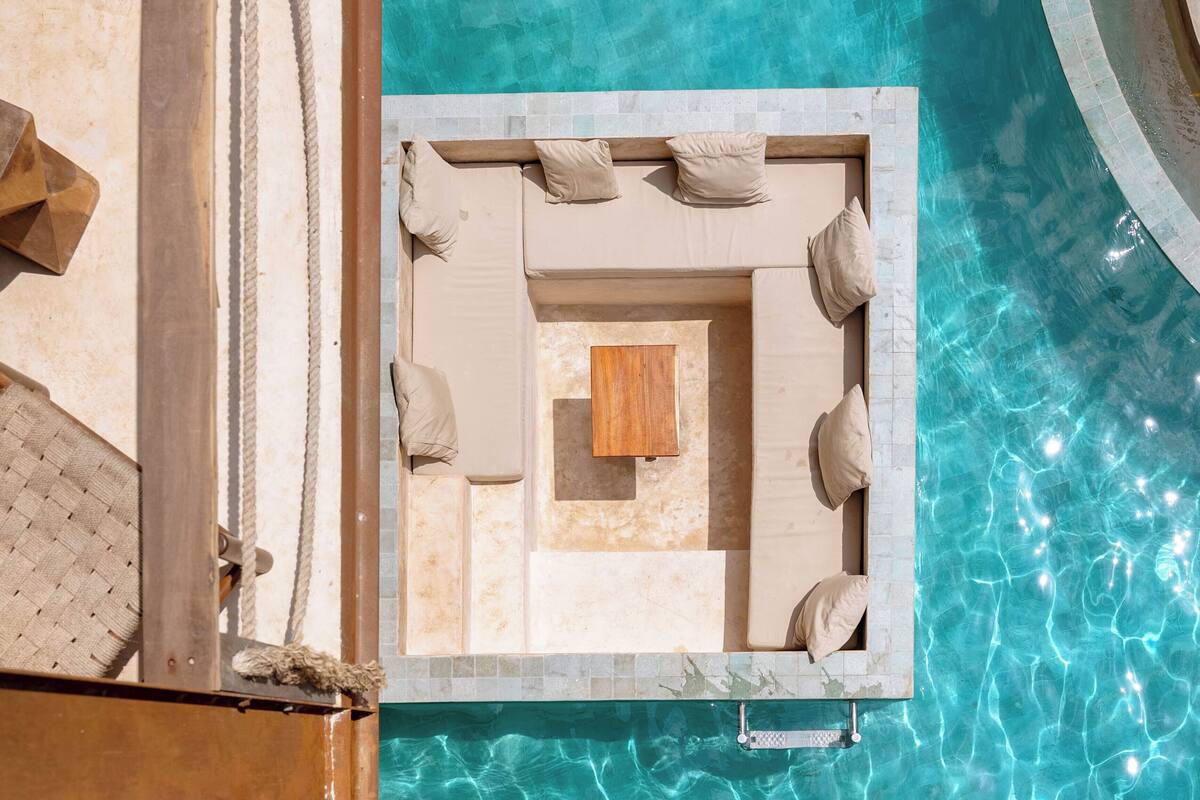 A poolside lounge area with a wooden table and beige cushions.