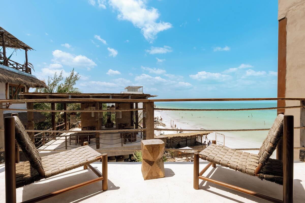 A beachside balcony with two wicker lounge chairs, a wooden table, and a thatched-roof structure.