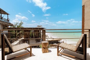 A beachside balcony with two wicker lounge chairs, a wooden table, and a thatched-roof structure. - Awa Holbox Hotel Boutique - Beach Front (Isla Holbox)