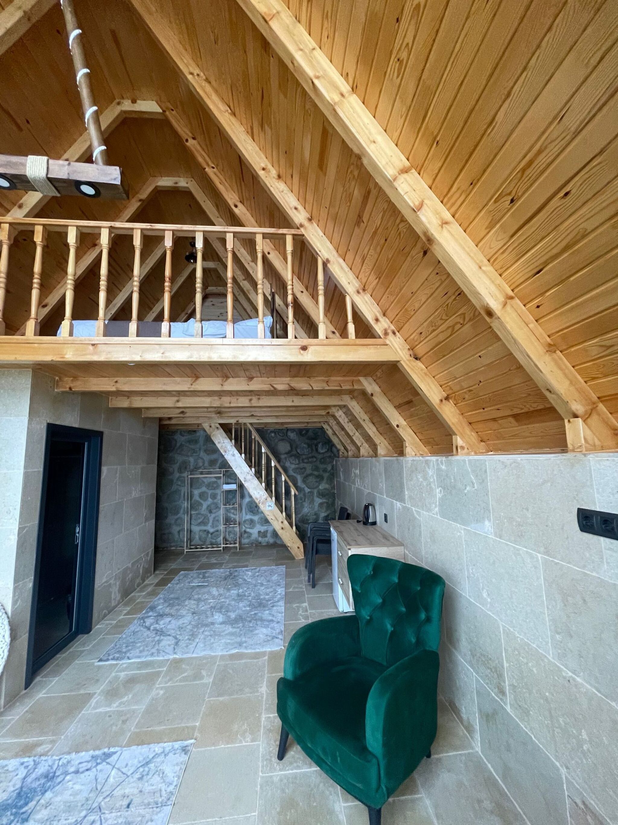 Ein geräumiger Innenraum mit hoher Holzbalkendecke, einer Wendeltreppe zu einer oberen Ebene, einem grünen Polsterstuhl und Steinwänden.