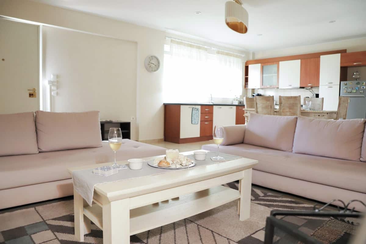 Uma sala de estar com sofá, mesa de centro com comida e bebidas, e cozinha ao fundo.