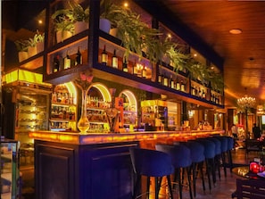 A bar area with multiple shelves of bottles, a counter with stools, and a decorative plant arrangement.