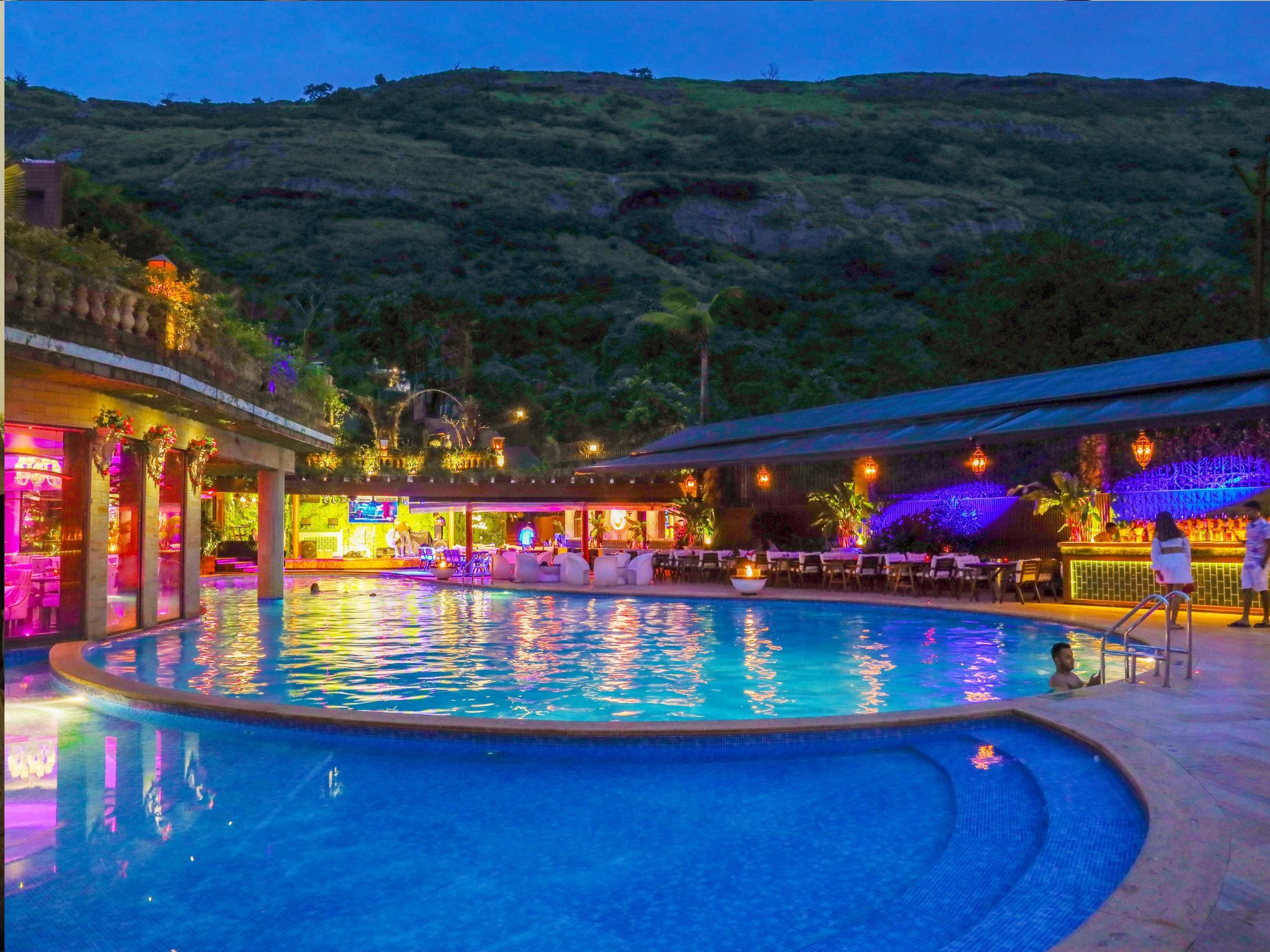 A swimming pool with a bar and seating area, surrounded by greenery and hills.