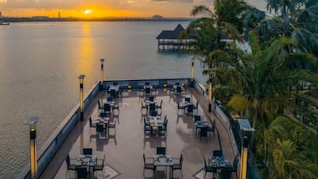 A waterfront restaurant with outdoor seating, a view of the sunset, and a distant overwater structure.