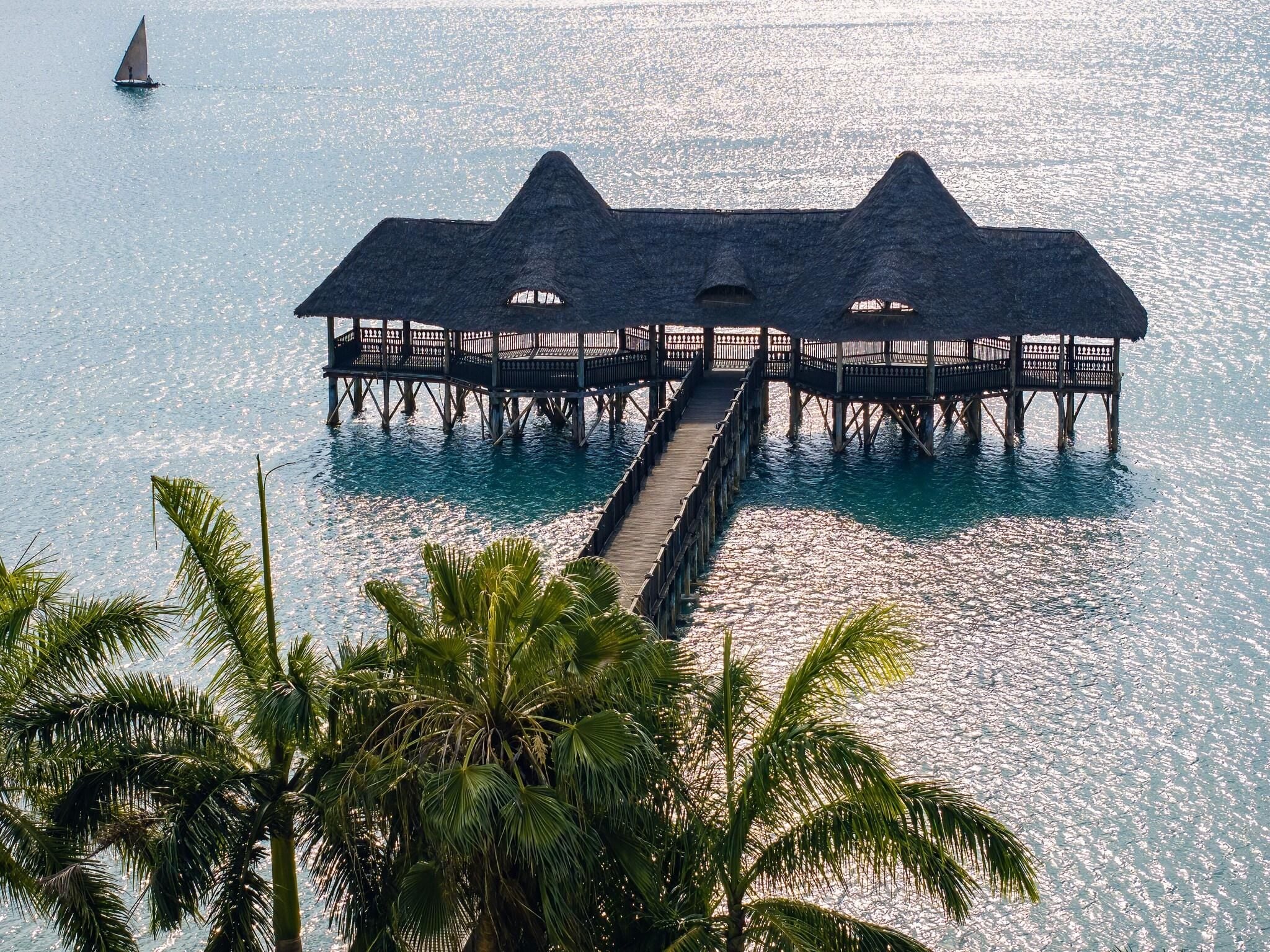 A wooden pier leading to a thatched-roof overwater structure with railings, surrounded by clear blue water and palm trees.