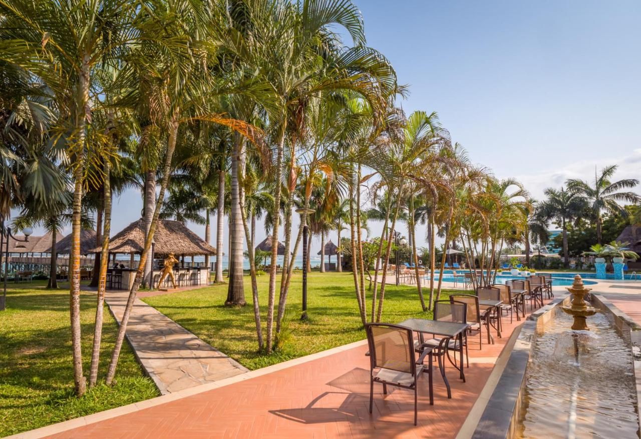 A resort with a pool, palm trees, outdoor seating, and a thatched-roof structure.