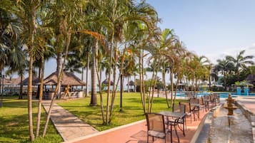 Un resort con piscina, palmeras, mobiliario exterior y una estructura de techo de paja.