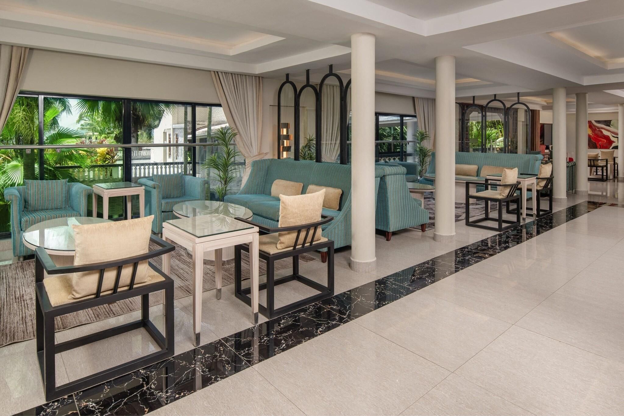 A spacious lobby with a seating area featuring sofas and chairs, large windows with curtains, and a view of greenery outside.