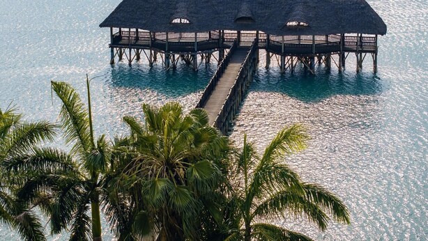A wooden pier leading to a thatched-roof overwater structure with railings, surrounded by clear blue water and palm trees. - Delta Hotels Marriott, Dar es Salaam (Dar es Salaam)