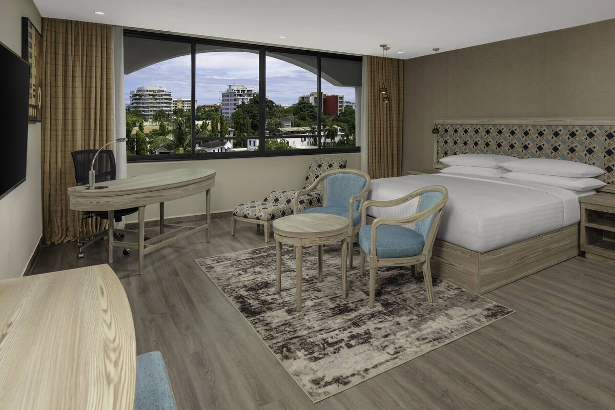 Habitación de hotel con una cama grande, dos sillas, un escritorio y vistas a edificios a través de un ventanal.