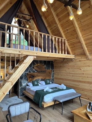 Interior em madeira com um quarto em mezanino, uma parede de pedra com um logotipo, uma cama, um banco e uma escada.