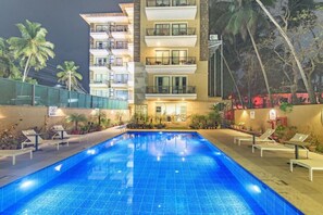 A swimming pool with lounge chairs, a multi-story building with balconies, and palm trees. - The Fern Residency, Calangute (Calangute)