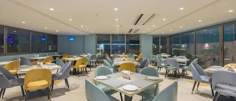 A spacious dining area with multiple tables and chairs, large windows, and a modern ceiling design.