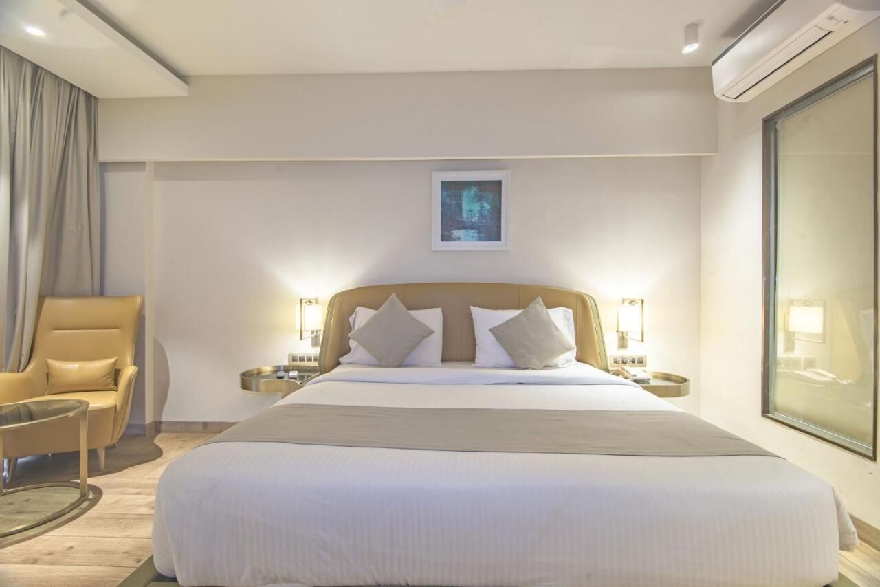 A hotel room with a large bed, a chair, bedside tables, a framed picture on the wall, and an air conditioning unit.