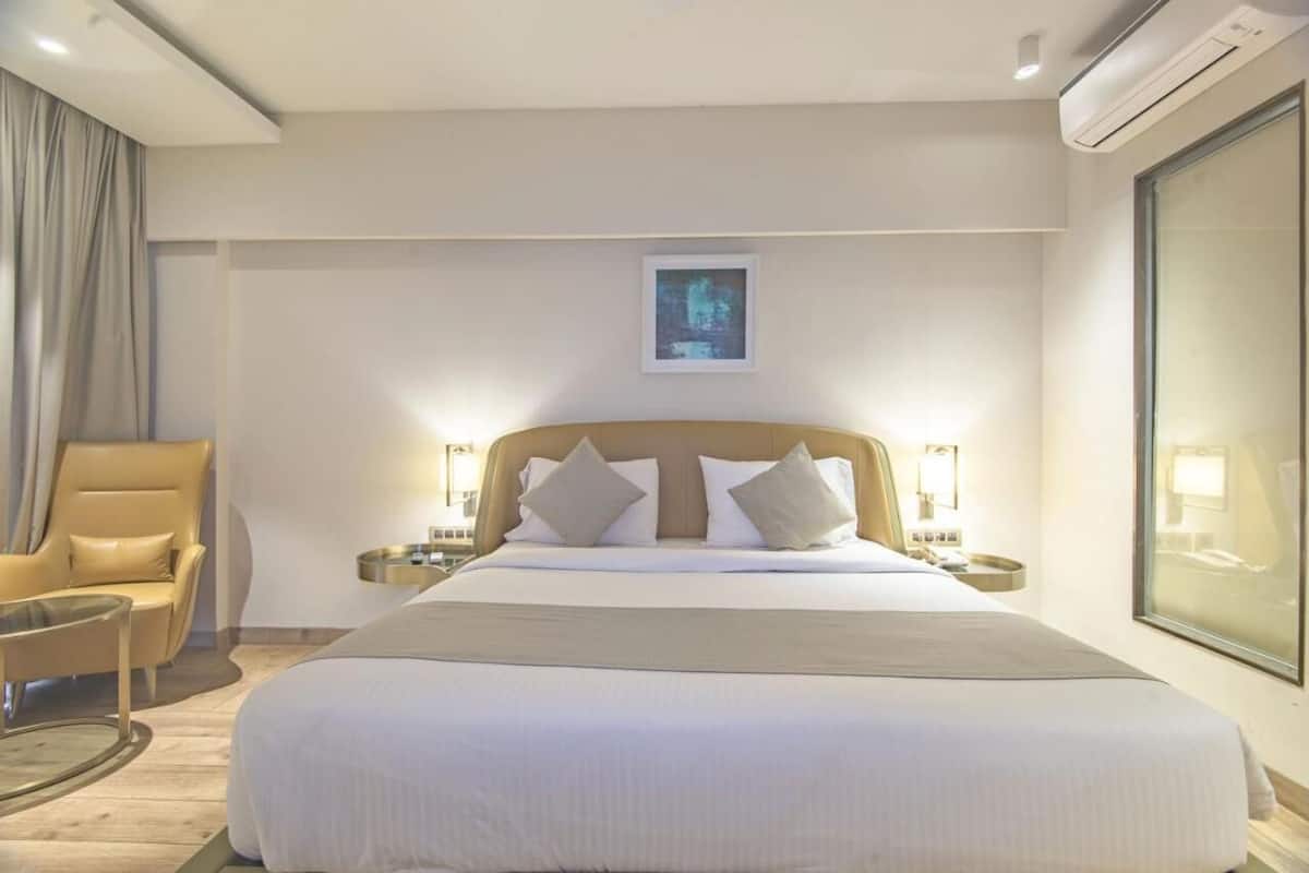 A hotel room with a large bed, a chair, bedside tables, a framed picture on the wall, and an air conditioning unit.