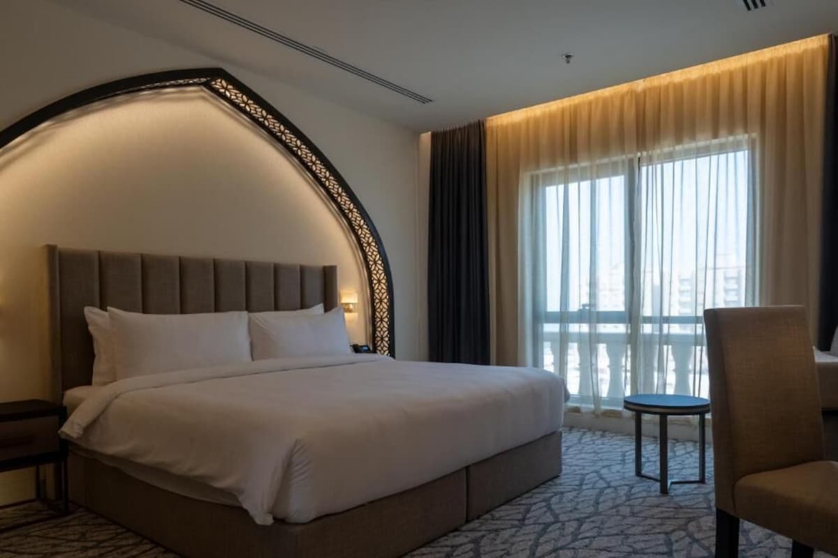 Ein Hotelzimmer mit einem großen Bett, einem Nachttisch, einem Sessel und Blick auf die Stadt durch große Fenster.