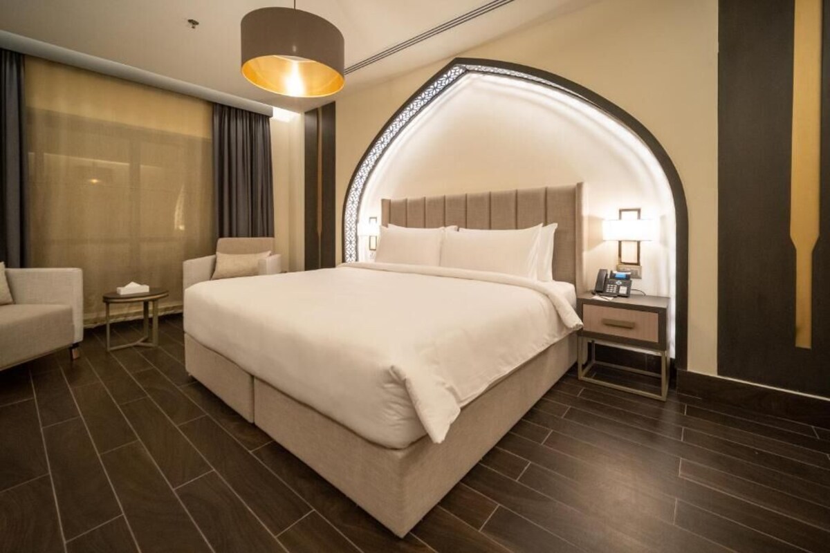 Ein Hotelzimmer mit einem großen Bett, einem Nachttisch mit Lampe, einem Sessel und einem großen Fenster mit Vorhängen.