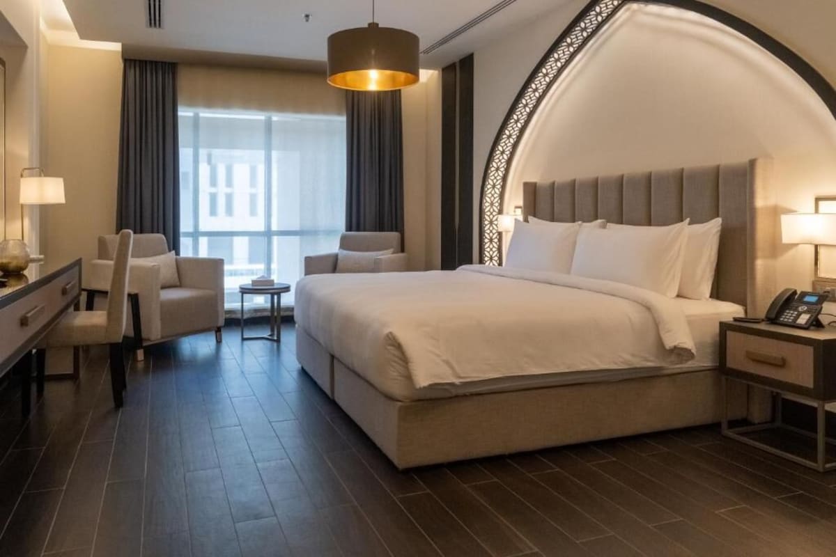Ein Hotelzimmer mit einem großen Bett, einem Schreibtisch, zwei Stühlen, einem kleinen Tisch und einem Telefon auf dem Nachttisch.