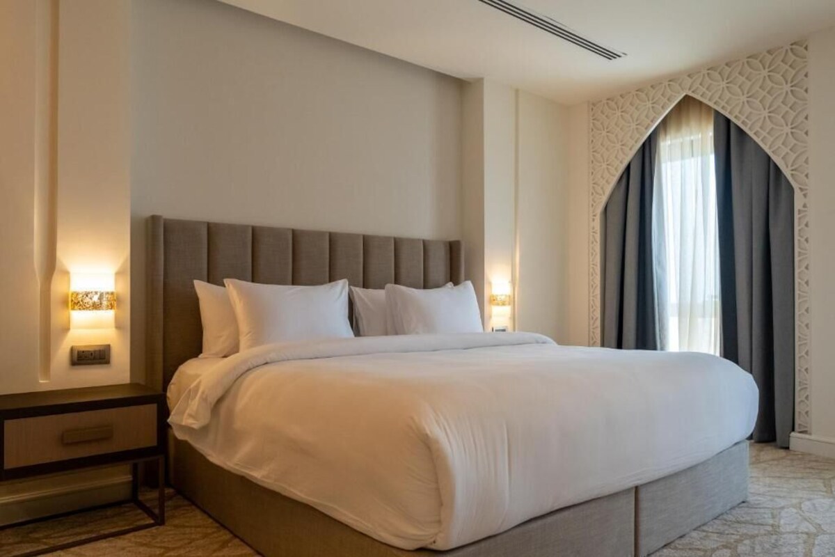 Ein Hotelzimmer mit einem großen Bett, einem Nachttisch, einer Wandlampe und einem Fenster mit Vorhängen.