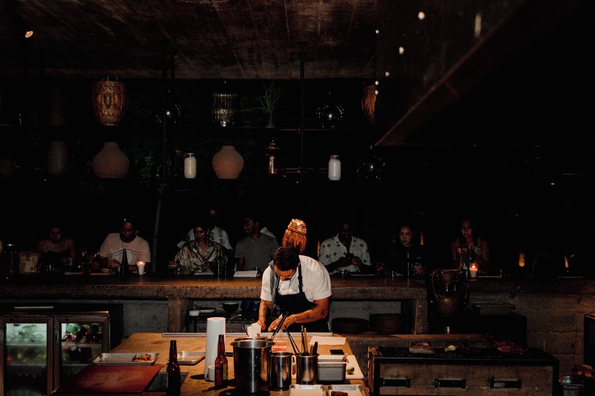 A chef preparing food at a counter in a dimly lit restaurant with patrons seated in the background.