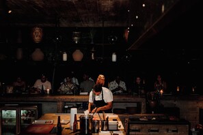 Un chef prépare des plats derrière un comptoir dans un restaurant à la lumière tamisée, tandis que des clients sont assis à l’arrière-plan.